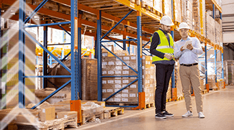 Two workers in hard hats and safety vests converse in a warehouse. They stand near tall shelves filled with boxes, discussing something on a tablet. The warehouse is well-lit and organized, with packaging materials on the floor.
