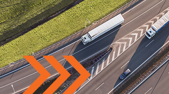 Aerial view of two white trucks and a car traveling on a highway divided by a patterned central barrier, with green grass on the sides. An orange arrow design overlays the bottom left corner of the image.