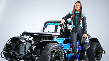 A person in a racing suit stands beside a black and blue race car, smiling. They hold a helmet in their right hand, resting their left hand on the car. The car displays the number 3 on its side.