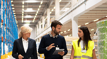 Three people walk through a warehouse. A man in the center holds a tablet, discussing with two women. One woman wears a suit, and the other a high-visibility vest. Shelving and products are visible in the background.
