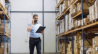A man in a light blue shirt holds a clipboard and talks on a phone while walking through a warehouse aisle. The shelves are stacked with brown cardboard boxes.