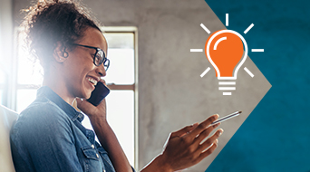 A person with curly hair and glasses smiles while talking on a phone and looking at a smartphone. There is an illustration of a lit light bulb on the right side of the image, suggesting a bright idea.