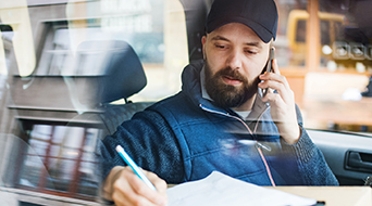 A man wearing a cap and a jacket is sitting inside a vehicle, talking on a cellphone. He is writing on a notepad with a pen. The focus is on his focused expression, and there is a reflection of the street in the window.