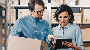 A man and a woman stand in a storage room with shelves filled with boxes. The man is holding a box, while the woman is pointing at a tablet. Both are wearing casual blue shirts and appear to be discussing something on the screen.