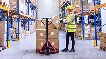 A woman in a warehouse wears a yellow hard hat and reflective vest as she operates a pallet jack, moving boxes. In this bustling small-business-shipping-hub, shelves filled with cardboard boxes line the walls. She pauses to check a label on one of the packages, ensuring precision.