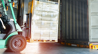 A forklift is loading a large pallet of goods into a shipping container inside a warehouse. The scene is lit by natural light coming from outside, highlighting the organized operation.