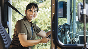 A delivery driver in a brown uniform, seated in a vehicle, smiles at the camera. Trees are visible outside the window, suggesting they are parked or stopped.
