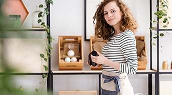 In a small-business hub, a person in a striped shirt and apron stands surrounded by shelves of wooden boxes and plants, carefully holding a small item.