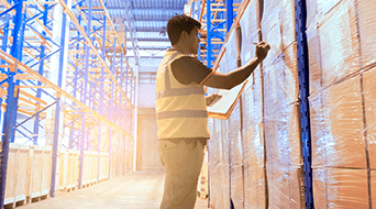 A person in a safety vest holds a clipboard and checks inventory in a warehouse. Tall shelves stacked with boxes line the aisle, and sunlight streams in from a high window, illuminating the space.