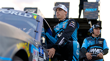 Race car driver kneeling beside a car, wearing a blue and black racing suit and cap. Another team member stands behind him wearing similar attire. The background features a trailer and logos.