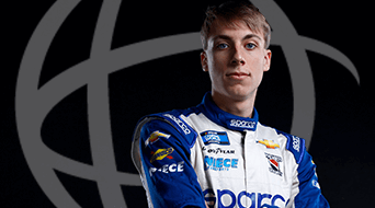A person in a blue and white racing suit stands with arms crossed against a dark background with a stylized globe logo. The suit features various sponsor logos, including Sparco and Chevrolet.