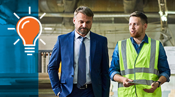 A man in a blue suit walks with a colleague in a safety vest and blue shirt inside an industrial setting, likely a bustling small-business-shipping-hub. The safety vest man gestures animatedly while talking. To the left, a light bulb graphic stands out on a vivid blue background.