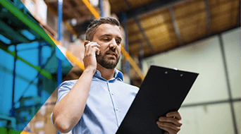 A man in a light blue shirt holds a clipboard and talks on the phone in a bustling small-business shipping hub. Shelves brimming with boxes fill the background, hinting at the warehouses lively operations.