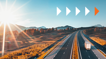 A truck is driving on an empty highway through a scenic landscape with mountains and trees under a clear blue sky. The sun is shining brightly, and there are white and an orange arrow pointing right above the scene.