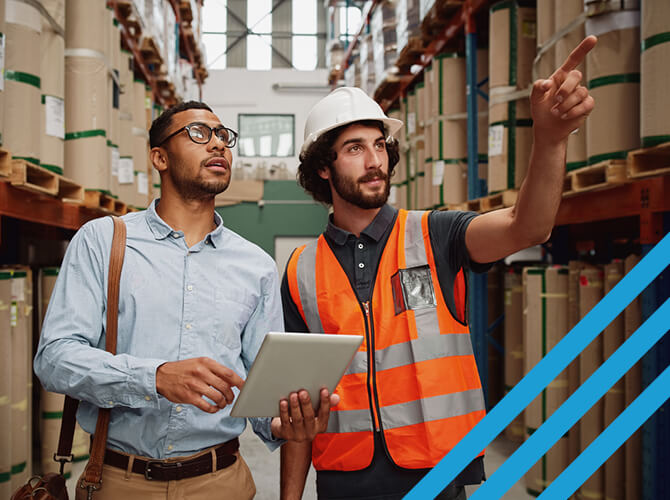 Two men in a warehouse, one with a tablet and glasses, the other in a hard hat and orange safety vest pointing forward. They stand amidst tall shelves with large rolls. Bright, well-lit industrial setting.