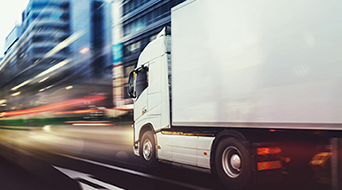 A white truck speeds through a blurred urban setting, embodying the energy of a small-business-shipping-hub. The cityscapes tall buildings are streaked with lights, highlighting the fast-paced movement and vibrant atmosphere.