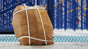 A large, crumpled, and tightly strapped cardboard package sits on the floor of a bustling small-business-shipping-hub, with blue metal shelving visible in the background.