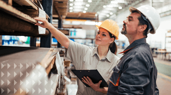 Two workers in a warehouse, both in hard hats. One holds a clipboard while pointing at a shelf, the other observes. Amidst industrial shelves and equipment of this small-business-shipping-hub, they laser-focus on inventory or inspection tasks to ensure efficient operations.