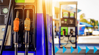 Close-up of two fuel pump nozzles at a gas station, with black and orange handles. A bright, sunlit background shows more fuel pumps and a partly cloudy sky, hinting at a busy station. Blue arrows are on the lower right corner.