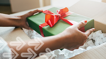 A person places a green gift box with a red ribbon into a cardboard box brimming with white packing peanuts. The scene evokes holiday shipping, where iconic arrows point from the lower left, suggesting movement and the excitement of sending gifts to loved ones.