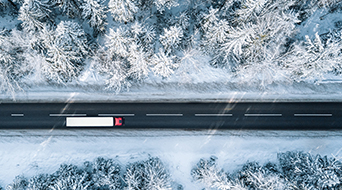 Aerial view of a red and white truck driving on a straight road surrounded by snow-covered trees and landscape. The scene is serene, with the snowy forest creating a stark contrast against the dark asphalt, capturing the essence of holiday shipping in winters tranquil embrace.