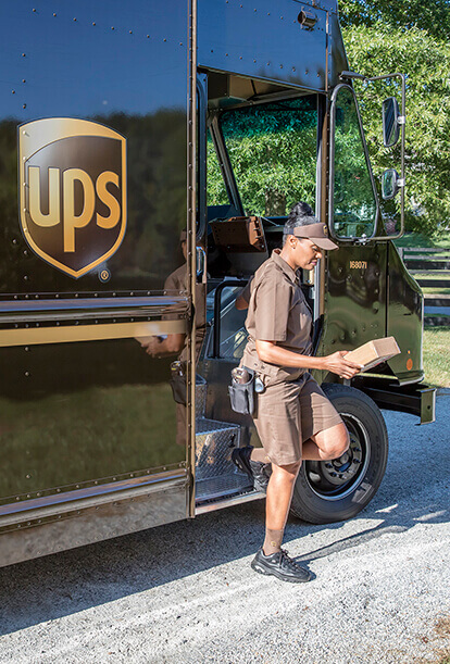 A UPS delivery person wearing a brown uniform examines a package while standing by an open truck door. The vehicle is parked on a sunny day, with green foliage in the background.