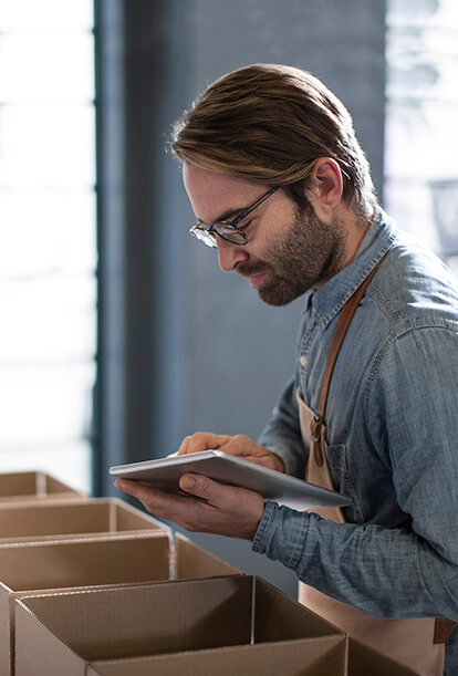 Man with glasses and a beard, wearing a denim shirt and apron, checking a clipboard next to a row of open cardboard boxes. He appears to be in a warehouse or workshop setting.