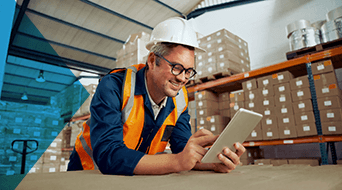 A person wearing a white hard hat and orange safety vest is smiling while using a tablet. They are standing in a warehouse with stacked cardboard boxes on shelves in the background.