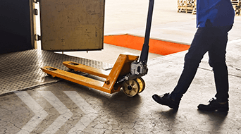 Person using a manual pallet jack to transport a wooden pallet through a warehouse door. The pallet jack is orange, and the floor has directional arrows marked. The person is wearing dark pants and shoes.