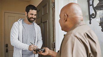 A man in a gray hoodie smiles while signing on a device held by another man in a brown jacket at a doorway, indicating an interaction at a small-business-shipping-hub.