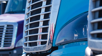 Close-up of the front grilles of two semi-trucks parked side by side at a small-business-shipping-hub. The truck in the foreground is blue, while the one partially visible in the background is purple. The focus is on the shiny chrome grille and reflective surfaces.