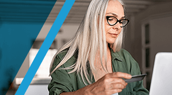 A woman with long white hair and glasses is holding a card while looking at a laptop screen. She is wearing a green shirt and is seated indoors. The image has a diagonal blue line on the left side.