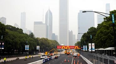 The image captures a Chicago street ready for a marathon, marked by orange cones and barriers. Skyscrapers, some shrouded in fog, rise in the background. Trees line the street as workers set up equipment, infusing the scene with an energy reminiscent of a bustling NASCAR event.