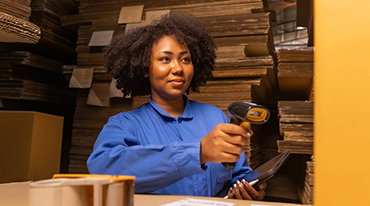 A woman with curly hair wearing a blue shirt is scanning a barcode with a handheld scanner in a warehouse filled with stacked cardboard boxes. She holds a tablet in her other hand.