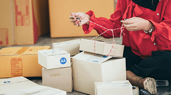 A person in a red shirt sits on the floor surrounded by cardboard boxes, tying red and white string. Some boxes are open, revealing packaging materials inside. The setting suggests a packing or shipping activity.