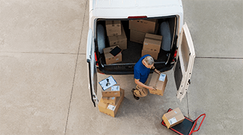 A person in a blue shirt is unloading boxes from the open back of a white van onto a flat surface. Various packages are scattered around, and a dolly is nearby. The situation appears to be a delivery or moving scenario.
