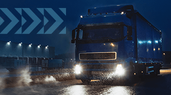 A large blue truck drives through a rainy, dimly lit industrial area at night. Its headlights illuminate the wet road. In the background, buildings and other vehicles are partially visible under cloudy skies.