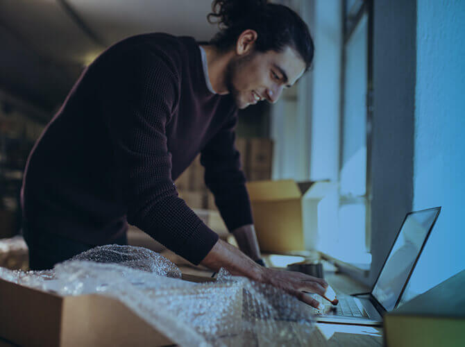 A person with long hair in a bun is standing by a desk, typing on a laptop. The person is surrounded by cardboard boxes and bubble wrap, suggesting a workspace related to packaging or shipping. The room is dimly lit.