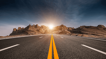 A straight highway stretches into the distance towards rocky hills under a dramatic, partly cloudy sky. The sun is setting or rising behind the hills, casting a warm glow over the landscape.