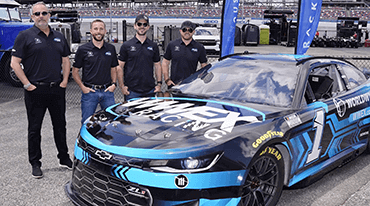 Four people stand next to a blue and black race car with the text Numex Racing on it. They are all wearing matching black shirts and sunglasses. The setting appears to be a racetrack with spectators in the background.