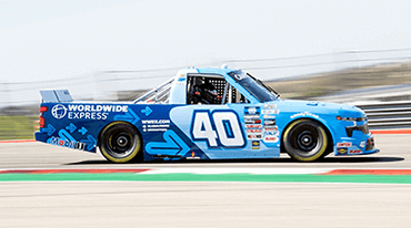 A blue race truck with the number 40 and Worldwide Express logo on the side speeds along a track. The image captures a side view of the vehicle in motion, showcasing sponsorship decals and blurred background indicating speed.