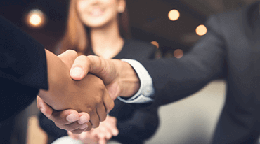 Two people are shaking hands in the foreground, symbolizing agreement or partnership. A blurred person in a suit is smiling in the background. The setting appears to be an indoor office space with soft lighting.