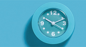 A round turquoise clock with white numbers and hands shows 10:10. The background is a matching turquoise shade, creating a monochromatic look. The clocks shadow is visible on the left side, adding depth to the image.