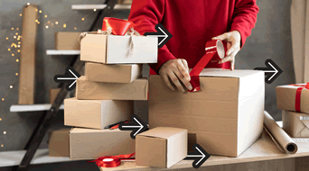A person in a red sweater is wrapping a large cardboard box with red ribbon on a table. Several other wrapped boxes and ribbons are stacked nearby. Black arrows point towards the boxes, and fairy lights are in the background.