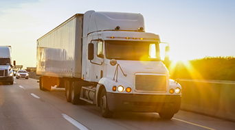 A white semi-truck with a trailer drives on a highway at sunset, aiming to manage freight costs efficiently. The sun creates a bright glare, casting long shadows on the road, as another truck passes in the opposite lane under a clear sky with a warm golden hue.