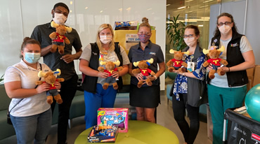 A group of six people wearing masks stand together indoors, holding stuffed animals and toys. They are dressed in business casual attire, and a small table in front of them has more toys. The background features a glass wall and office furniture.
