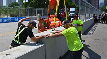 Workers in safety vests and hats secure concrete barriers on a street as if prepping for a NASCAR event. A cherry picker is in the background, and nearby stands a Blue Shield sign along the fence. Trees and city buildings frame this bustling scene, reminiscent of Chicagos dynamic energy.