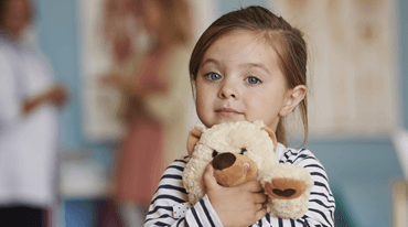 A young girl with brown hair holds a teddy bear in her hands, looking directly at the camera. She is wearing a striped shirt. The background shows blurred figures of adults and a light blue wall.