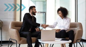 A man and woman in business attire are shaking hands, sitting on a couch in an office setting. A laptop and a notepad are on the round table between them. An arrow graphic is on the left side of the image.