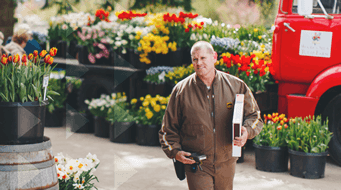 A delivery person in a brown uniform walks in front of a red truck with colorful flowers in large pots. They carry a small package and device, surrounded by blooming tulips and daffodils at an outdoor market.